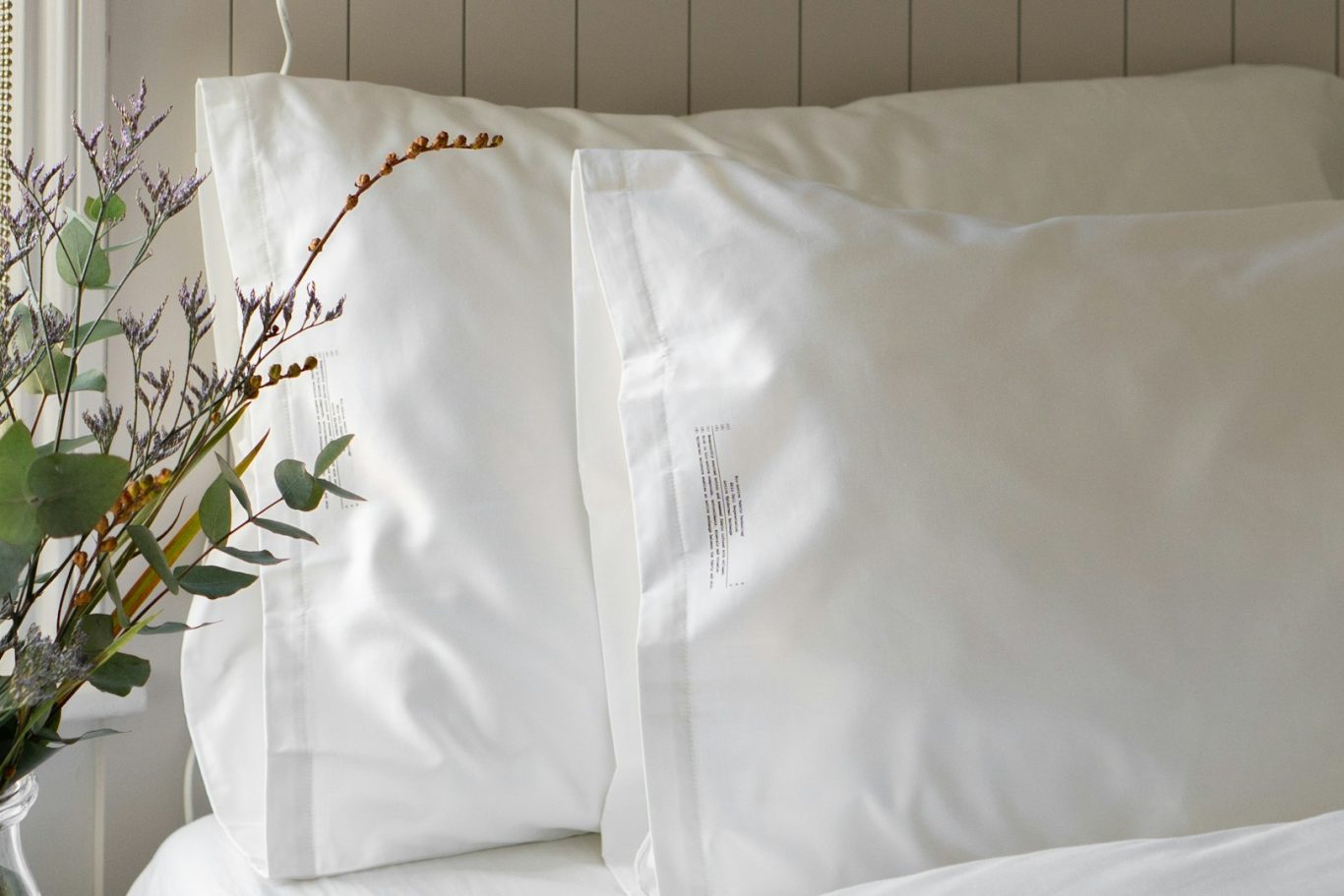 PACK REVALORIZA Dos almohadas blancas sobre una cama con un ramo de flores en un jarrón.